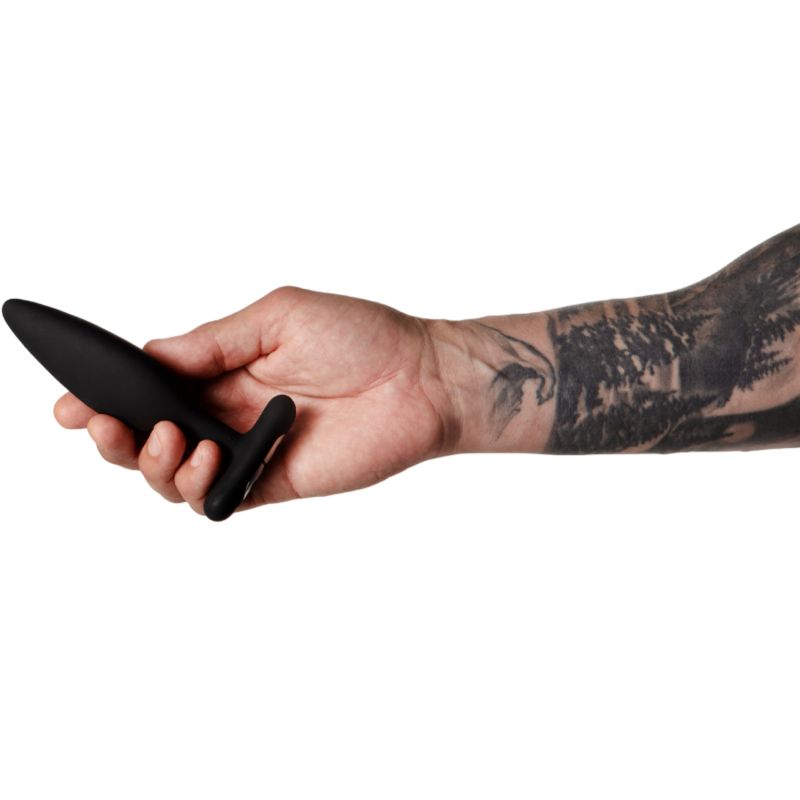 Hand hält einen schwarzen Analplug. Arm mit Tätowierung. Weißer Hintergrund.