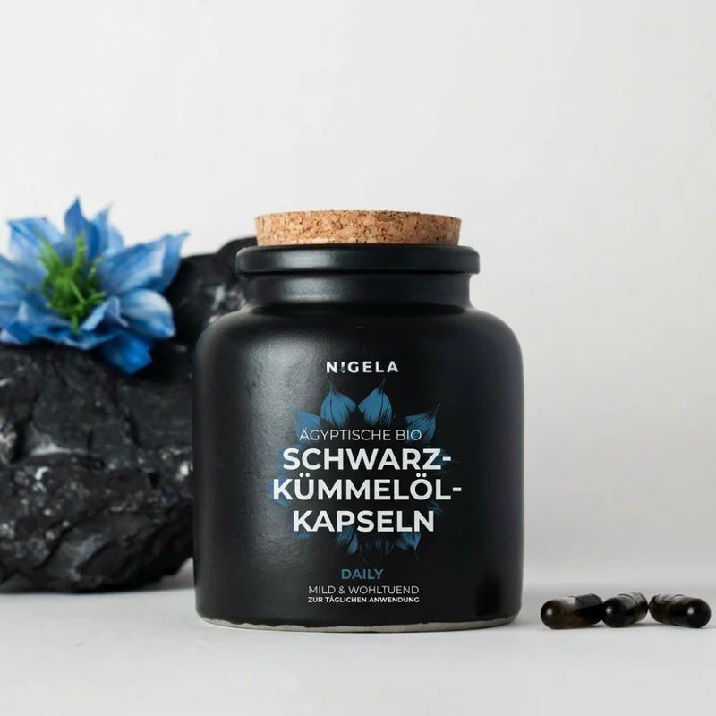Schwarzkümmelöl Kapseln in schwarzer Keramikdose mit Korkverschluss, daneben drei Kapseln und eine blaue Blüte.
