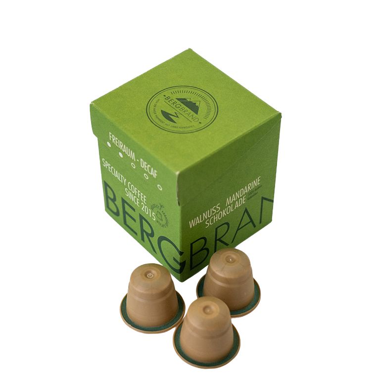 Grüne Verpackung mit BERGBRAND-Logo und Kaffeekapseln. Text: Freiraum-Decaf, Specialty Coffee, Walnuss Mandarine Schokolade.
