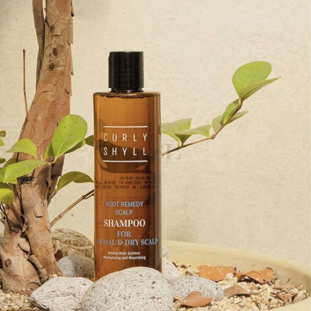 Braune Shampoo-Flasche mit schwarzem Deckel. Aufschrift: CURLY SHYLL, Root Remedy Scalp Shampoo, für normale und trockene Kopfhaut. Umgeben von Pflanzen.