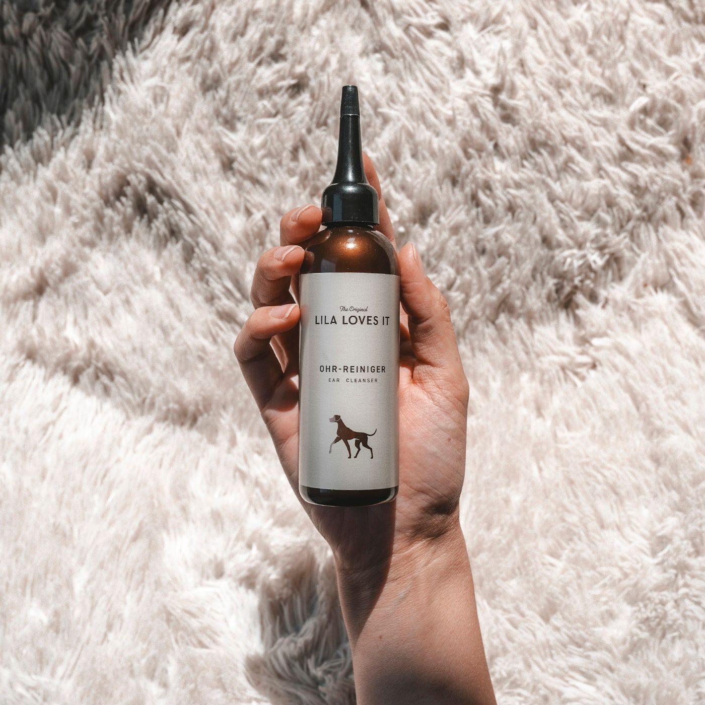 Braune Flasche mit schwarzem Applikator. Etikett mit "LILA LOVES IT" und "Ohr-Reiniger". Hand hält die Flasche.