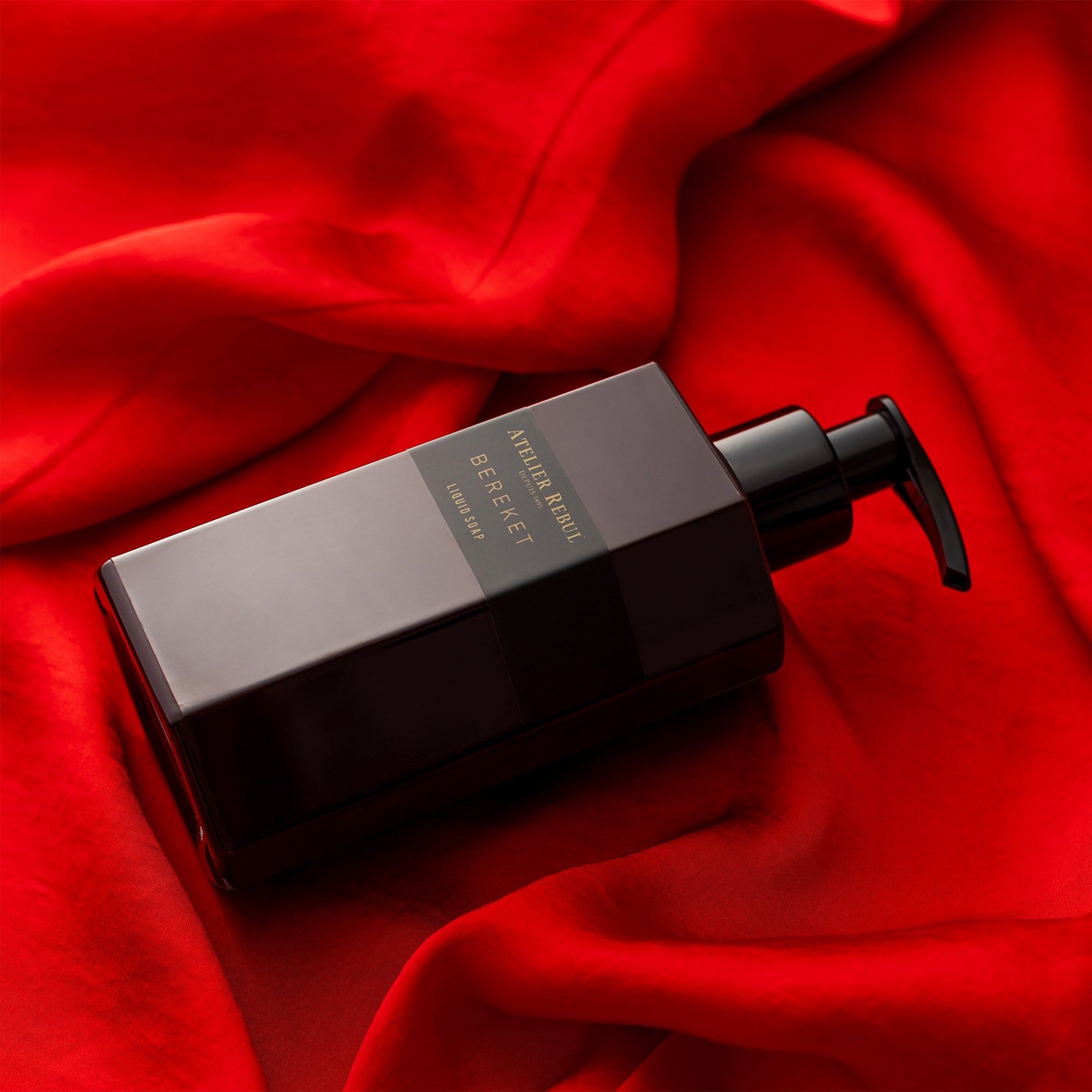 Bordeauxfarbene Flüssigseife mit schwarzem Pumpkopf liegt auf rotem Seidenstoff. Aufschrift: Atelier Rebul Bereket.