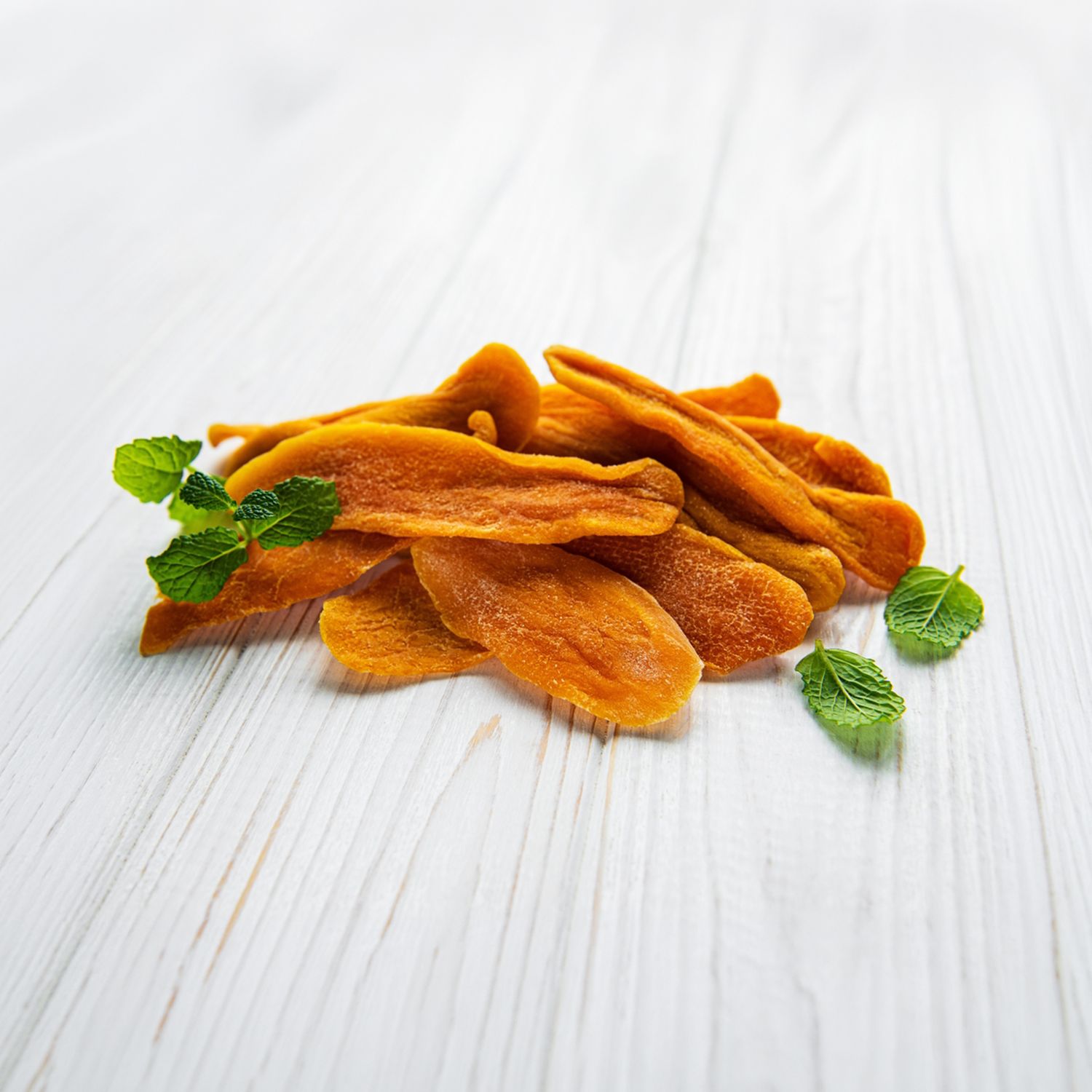 Getrocknete Mangoscheiben mit Minzblättern auf einer weißen Holzoberfläche.