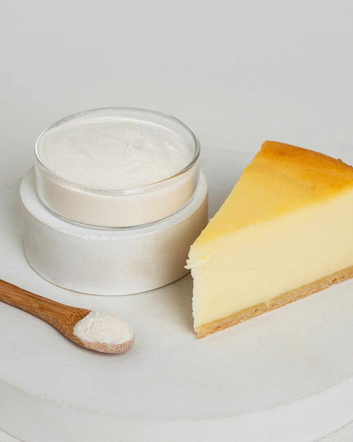 Xanthan Gum Pulver in einem Glasbehälter neben einem Stück Käsekuchen und einem Holzlöffel.