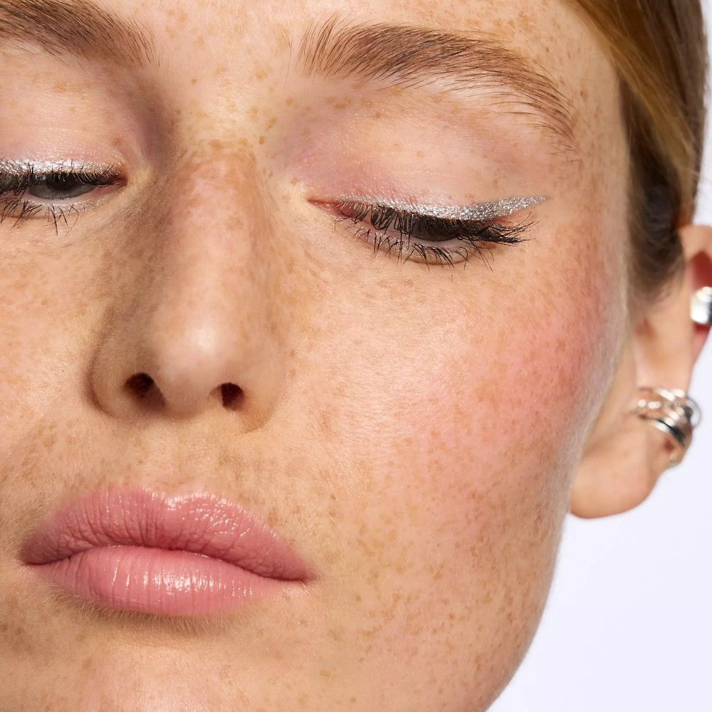 Nahaufnahme eines Gesichts mit silbernem Eyeliner. Geschlossene Augen, rosige Wangen und Lippen.