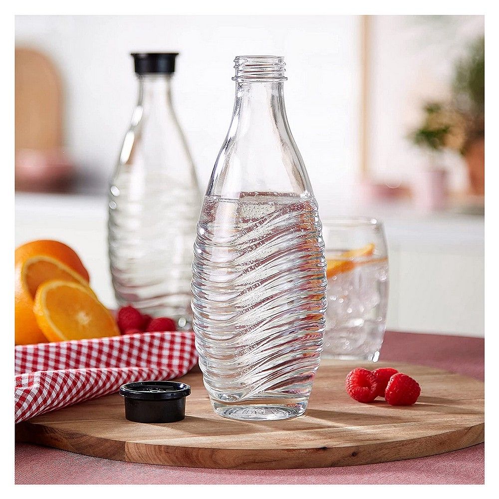 Eine SodaStream Glasflasche mit schwarzem Deckel, gefüllt mit sprudelndem Wasser, auf einem Holzteller mit Früchten.