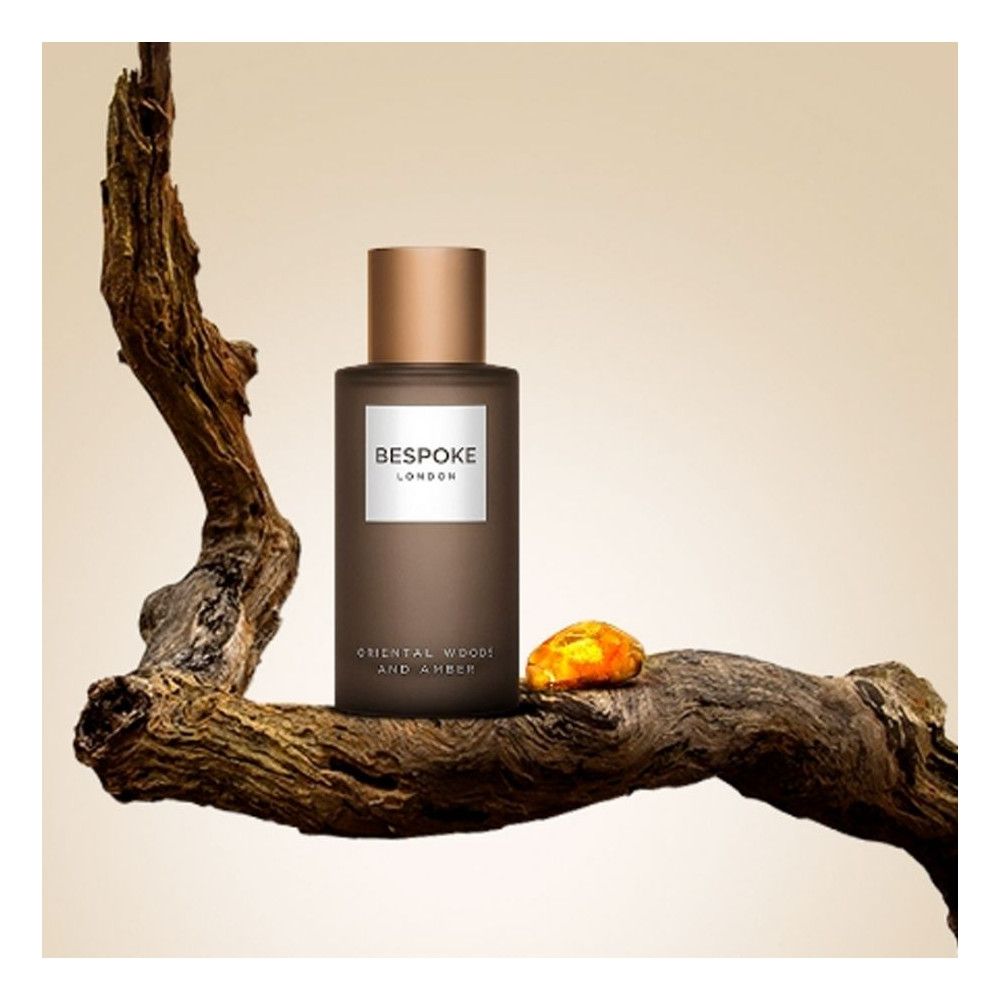 Parfümflakon mit goldfarbenem Deckel. Aufschrift: Bespoke London, Orientalische Hölzer und Amber. Liegt auf einem Ast und einem Bernstein.