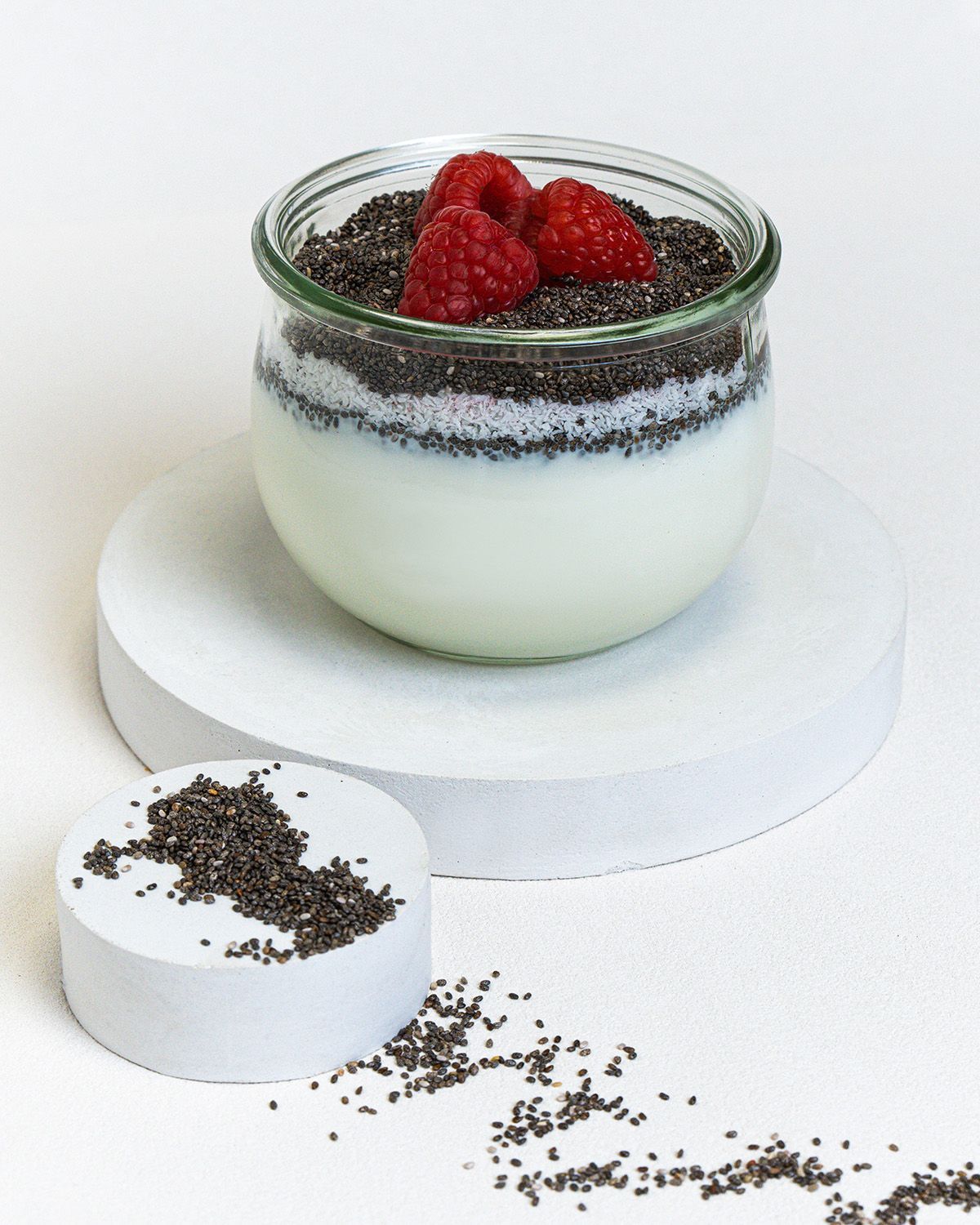 Glas mit Joghurt, Chia Samen und Himbeeren. Auf weißem Untergrund. Einzelne Samen verstreut.