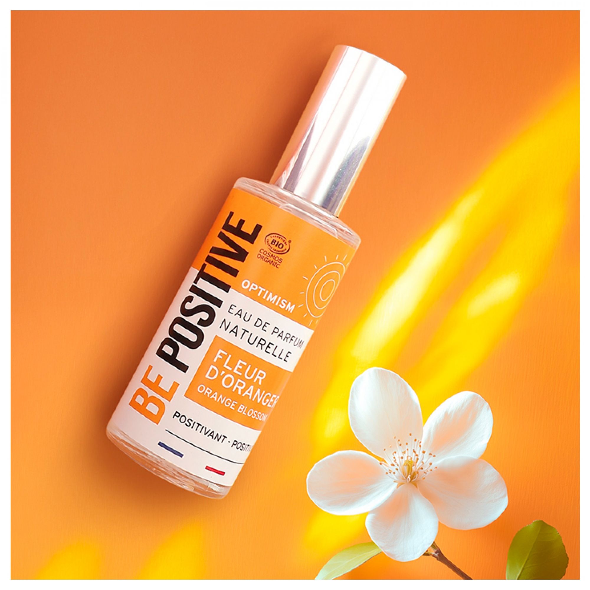 Acorelle Be Positive Eau de Parfum Optimism Fleur d'Oranger neben einer Orangenblüte auf orangefarbenem Hintergrund.