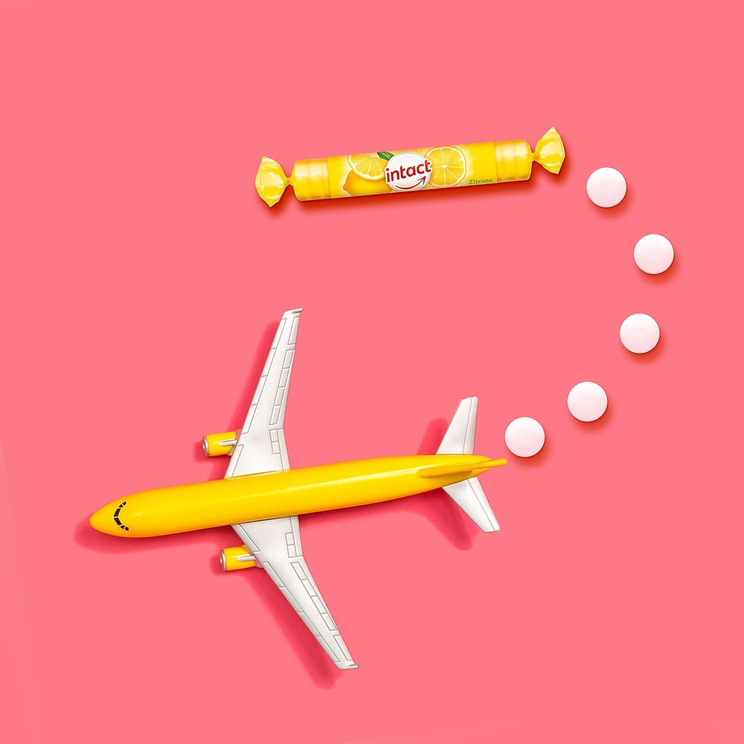Gelbes Spielzeugflugzeug und eine Rolle Traubenzucker. Daneben weiße Tabletten, die eine Flugbahn darstellen. Hintergrund: rosa.