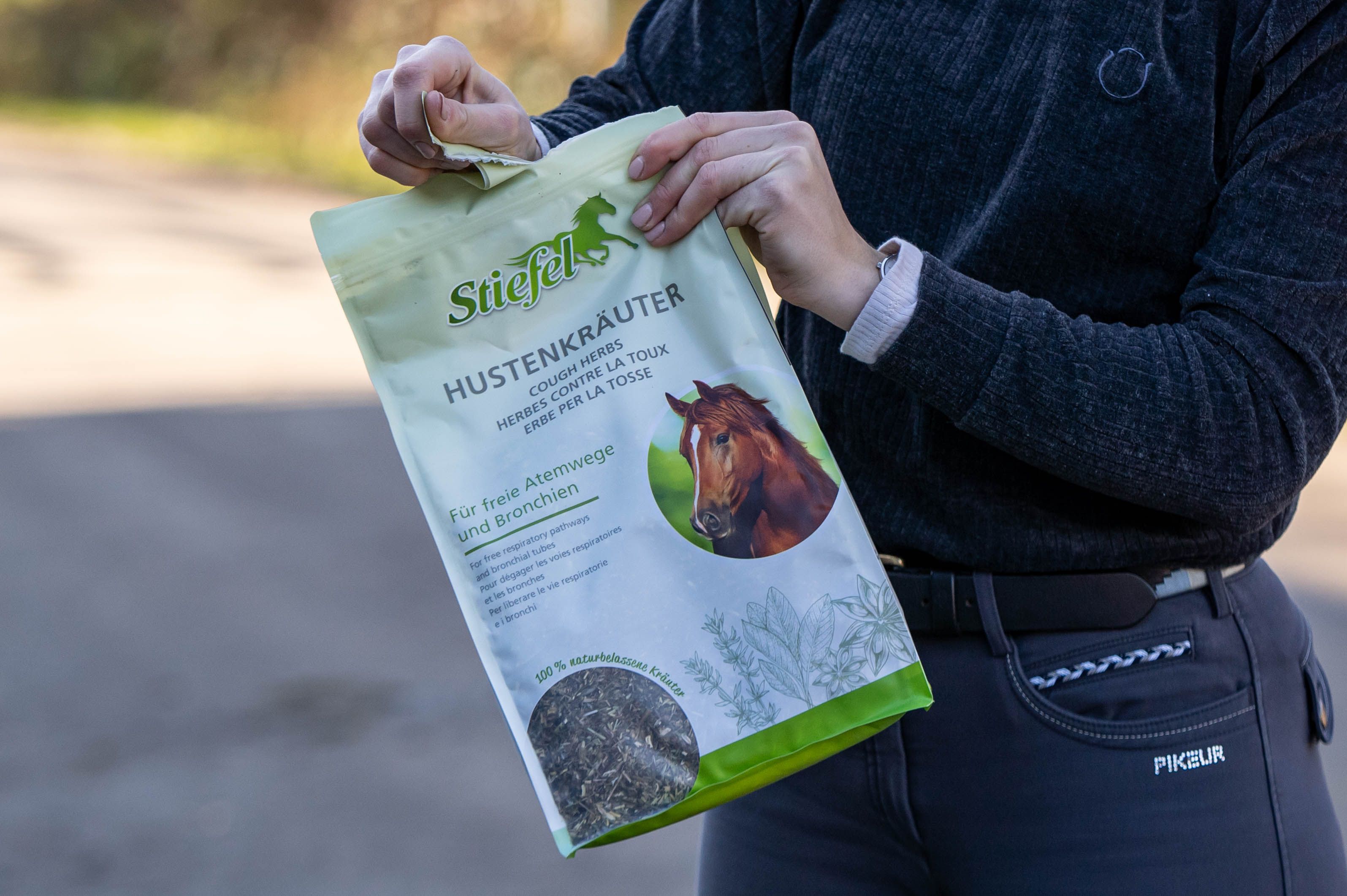 Person öffnet einen Stiefel Hustenkräuter-Beutel. Der Beutel zeigt das Produkt, den Markennamen und einen Pferdekopf. Für freie Atemwege und Bronchien.
