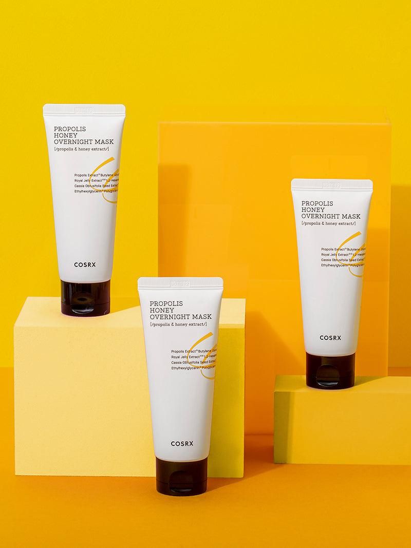 Drei weiße Tuben mit schwarzem Deckel. Aufschrift: PROPOLIS HONEY OVERNIGHT MASK. Marke: COSRX. Auf gelbem Hintergrund.