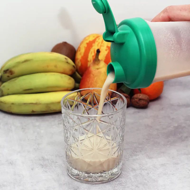 Getränk wird in ein Glas gegossen. Shaker mit grünem Deckel. Früchte im Hintergrund: Bananen, Birne, Orange, Kiwi.