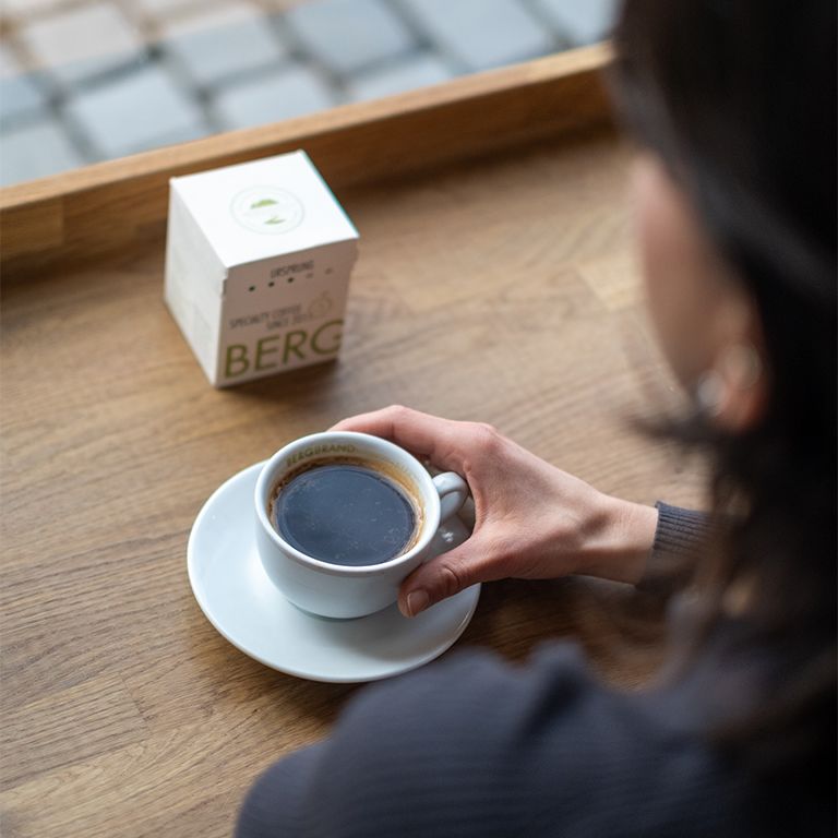 Kaffeetasse auf Untertasse. Daneben Verpackung. Aufschrift: BERGBRAND, Ursprung, Specialty Coffee. Person hält Tasse.
