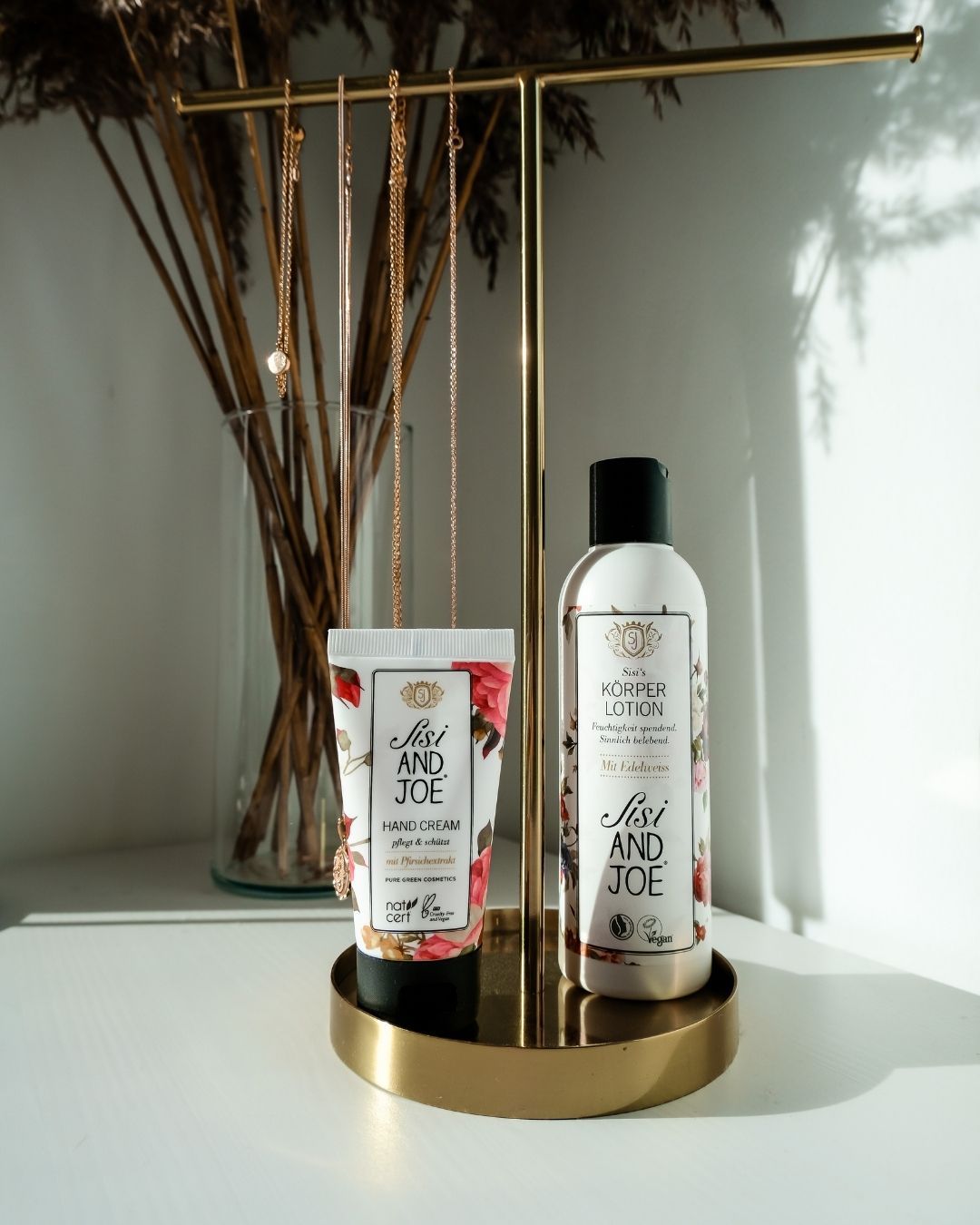 Handcreme und Körperlotion von Sisi and Joe. Beide Produkte stehen auf einem goldenen Ständer. Die Flaschen sind mit Blumen verziert.