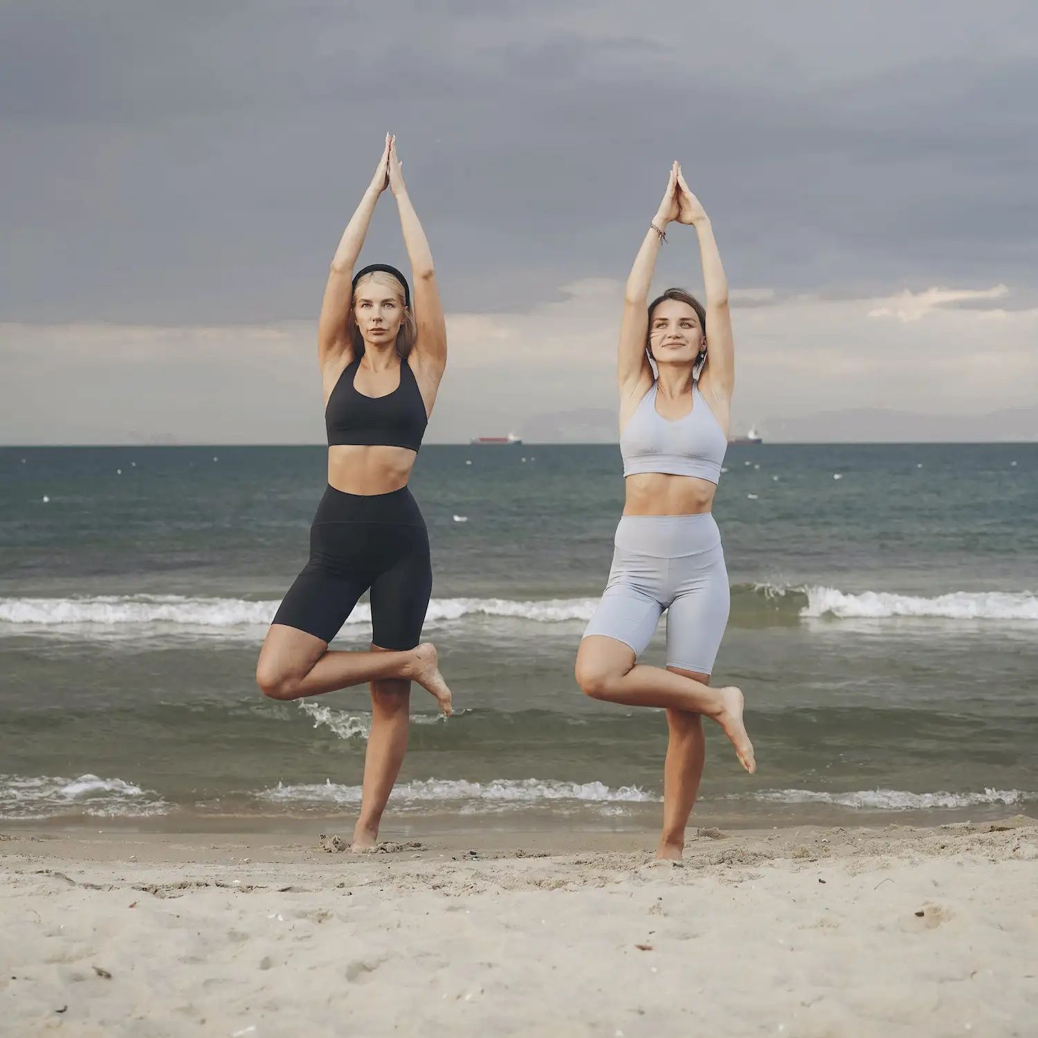 Zwei Frauen in Sportkleidung stehen am Strand in Yoga-Pose. Meer und Himmel im Hintergrund.