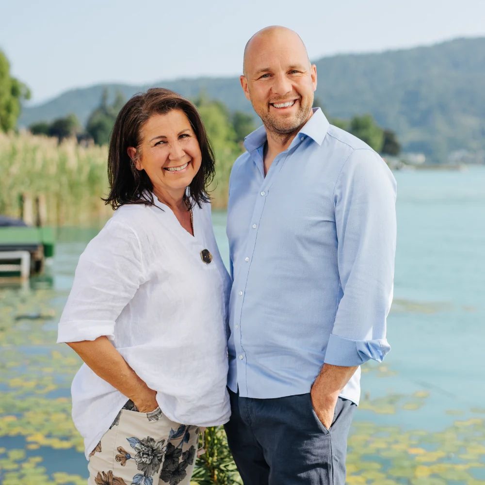Ein Mann und eine Frau stehen lächelnd nebeneinander am Ufer eines Sees.