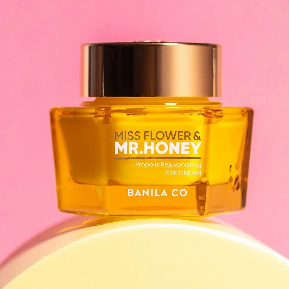 Goldfarbener Tiegel Augencreme. Aufschrift: Miss Flower & Mr. Honey, Propolis Rejuvenating Eye Cream, BANILA CO. Auf rosa Untergrund.