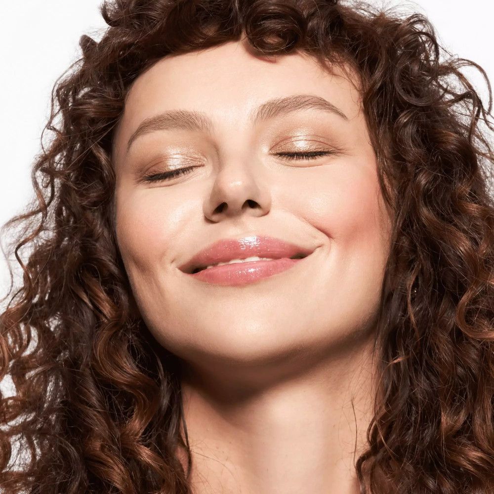 Nahaufnahme einer Frau mit geschlossenen Augen. Sie trägt hellen Lidschatten und rosa Lippenstift. Lockiges, braunes Haar.