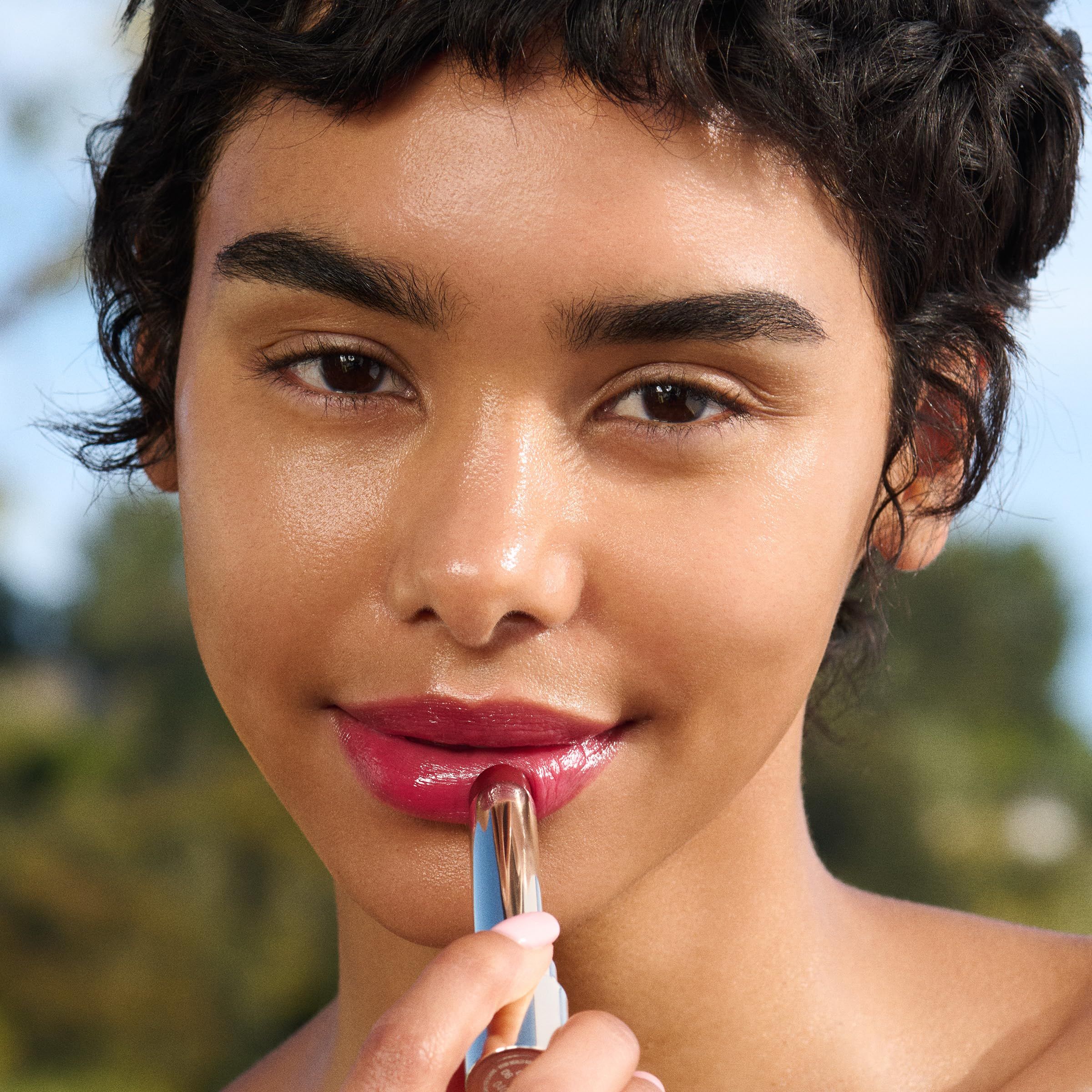 Person trägt Lip Oil Balm auf. Person mit dunklen Haaren und Haut. Produkt in Anwendung.