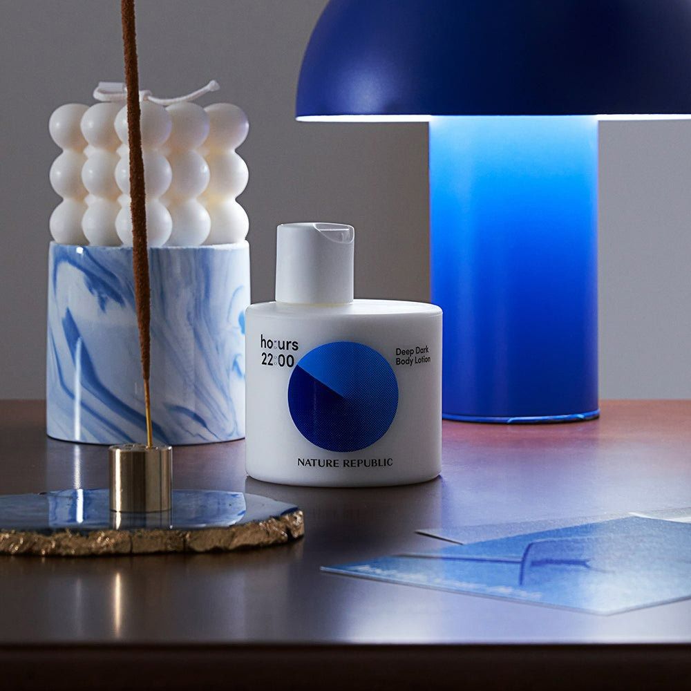 Weiße Flasche mit blauem Kreis, neben Kerze und Lampe. Text: ho:urs 22:00, Deep Dark Body Lotion, NATURE REPUBLIC.