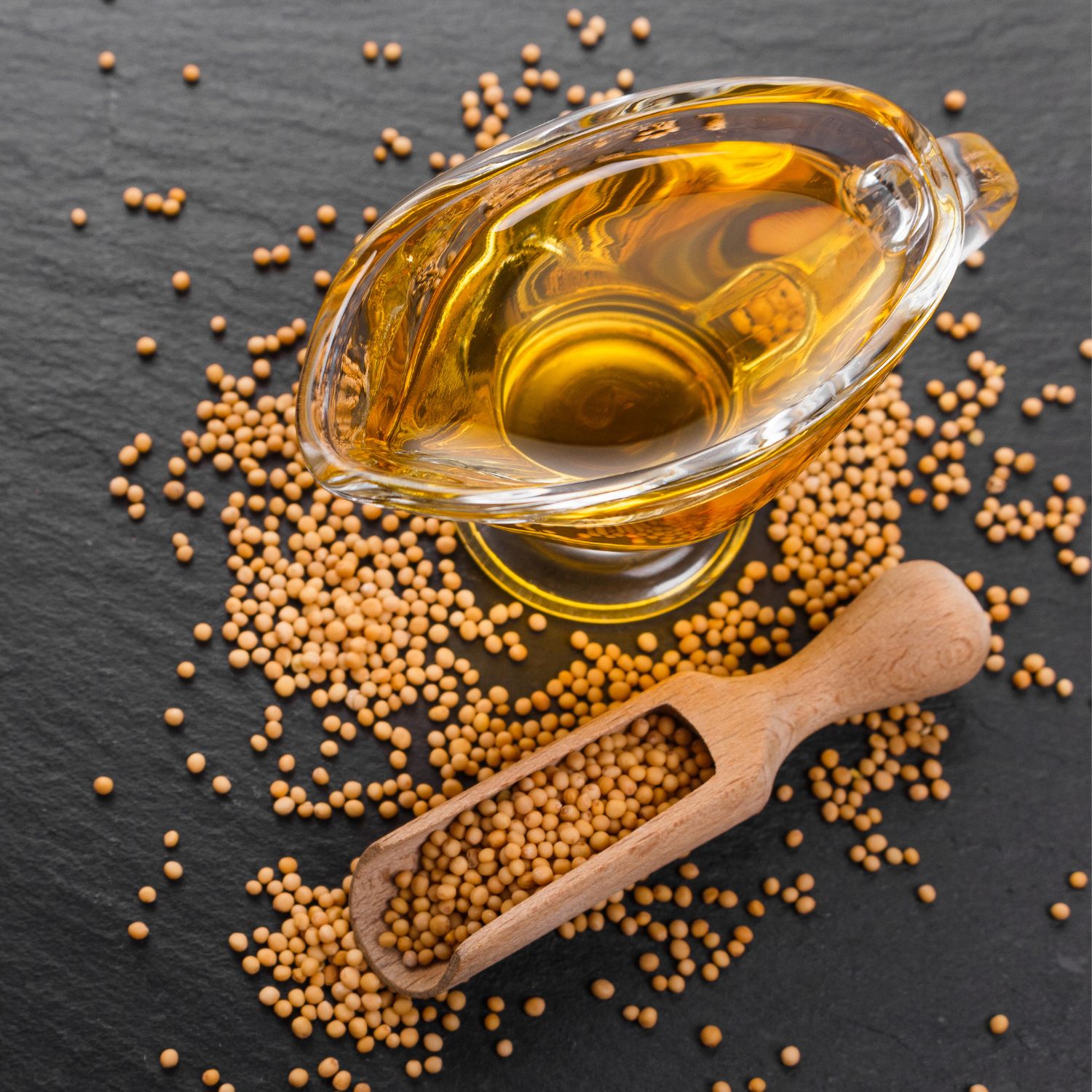 Senf in Glas-Sauciere, umgeben von Senfkörnern und Holzschaufel. Auf schwarzem Untergrund.