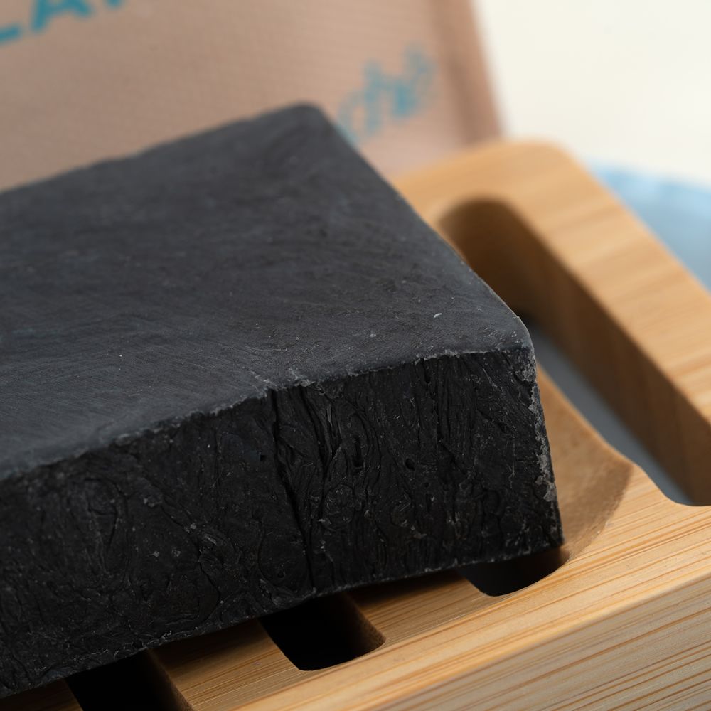 Schwarze Seife auf Holzseifenschale. Seife rechteckig. Hintergrund: hellblaues Tuch.