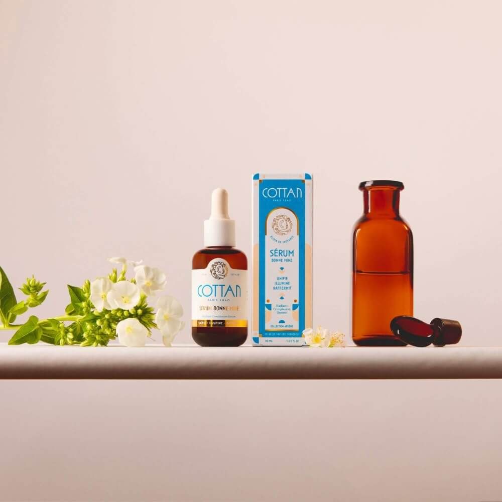 Serumflasche und Schachtel mit Aufschrift COTTAN, Serum Bonne Mine. Daneben eine leere Flasche und Blumen.