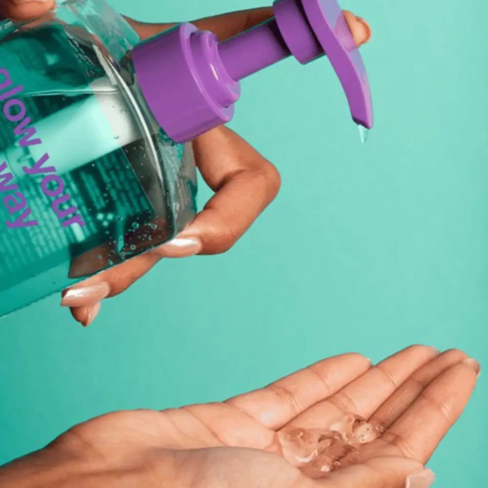 Hand gießt Flüssigkeit aus einer Flasche mit lila Pumpe in eine andere Hand. Auf der Flasche steht "glow your own way".