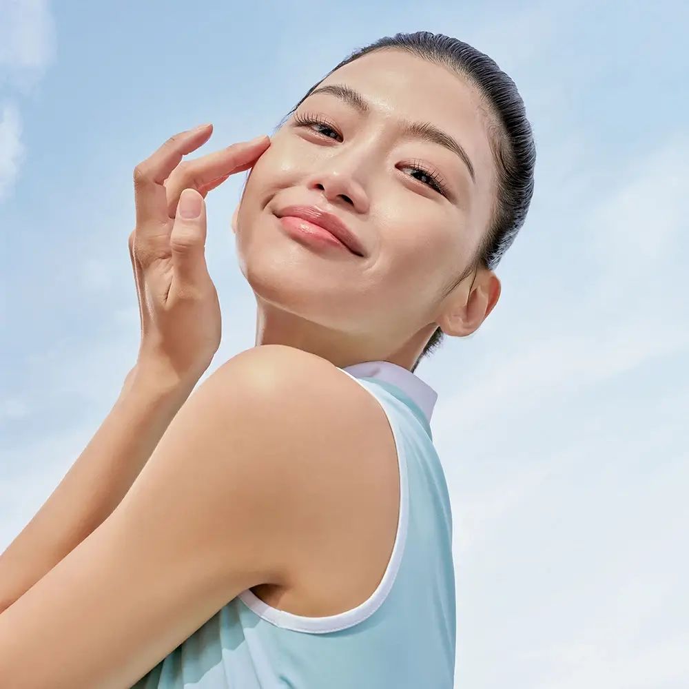 Junge Frau berührt sanft ihr Gesicht. Blauer Himmel im Hintergrund. Sie lächelt.