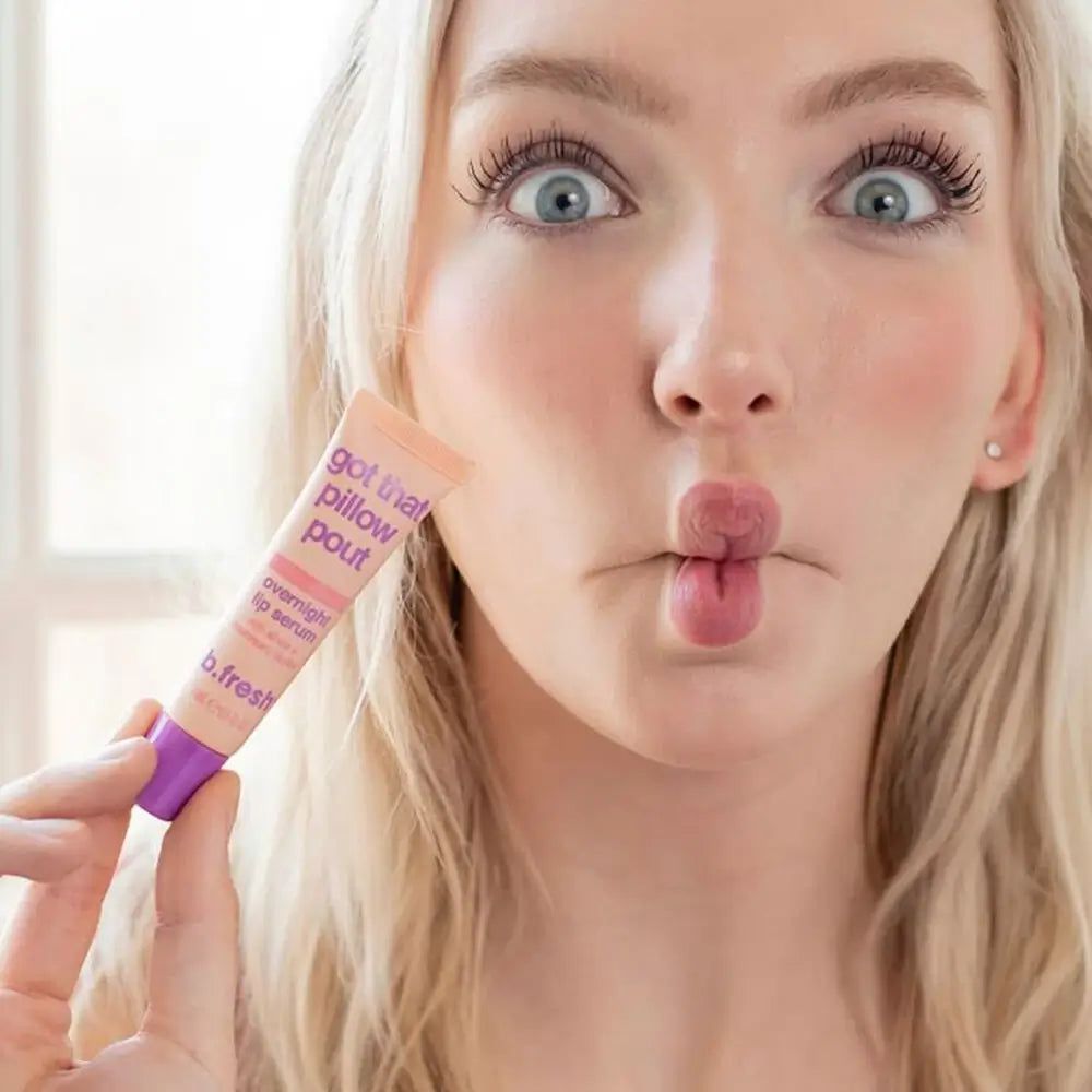 Frau hält Lippen-Serum. Auf dem Produkt steht: Got That Pillow Pout. Die Marke ist b.fresh. Das Produkt ist in einer Tube.