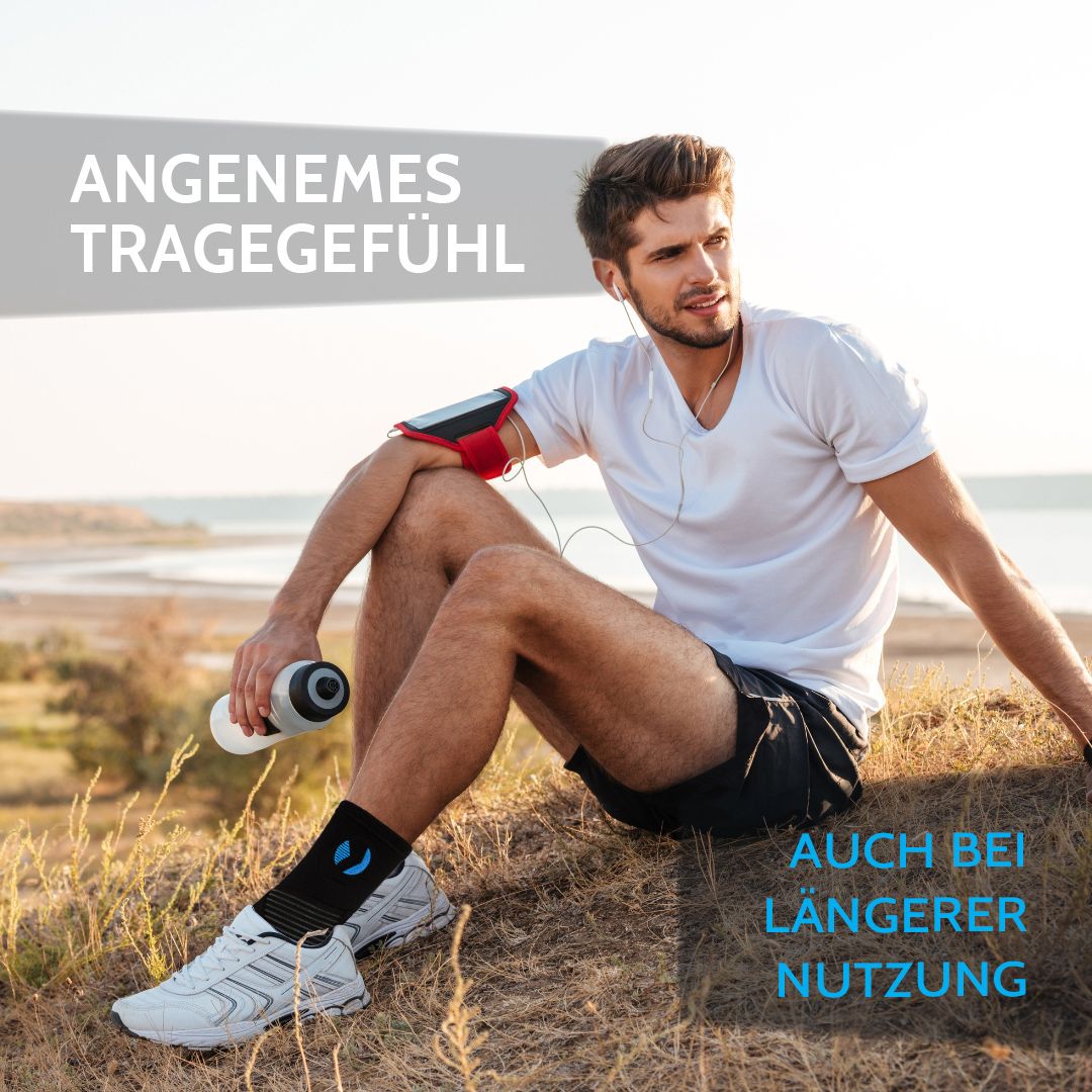 Mann sitzt mit Sprunggelenkbandage am Strand. Schwarze Bandage mit blauem Logo. Angenehmes Tragegefühl, auch bei längerer Nutzung.