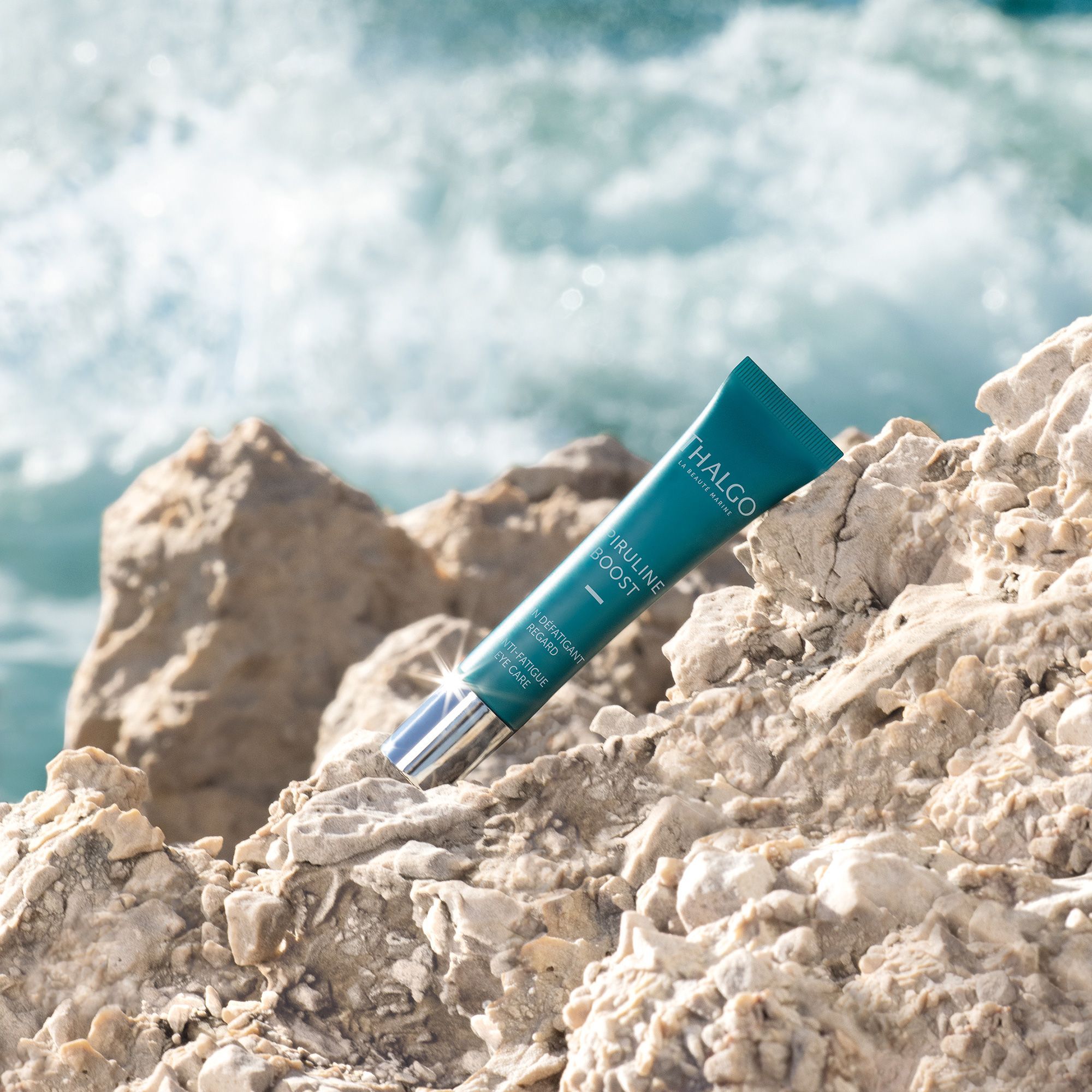 Türkisfarbene Tube auf Felsen. Aufschrift: THALGO, Spiruline Boost, Anti-Fatigue Eye Care. Hintergrund: Wasser.