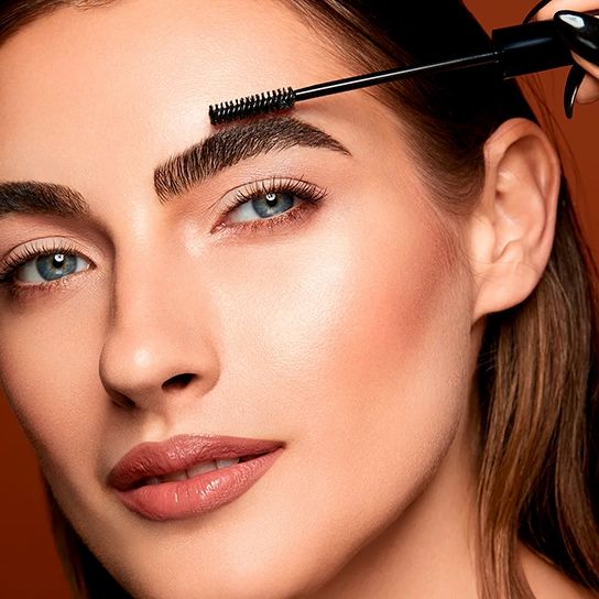 Frau mit Augenbrauen, die mit einem schwarzen Bürstchen bearbeitet werden. Braune Augenbrauen, blaue Augen, brauner Hintergrund.