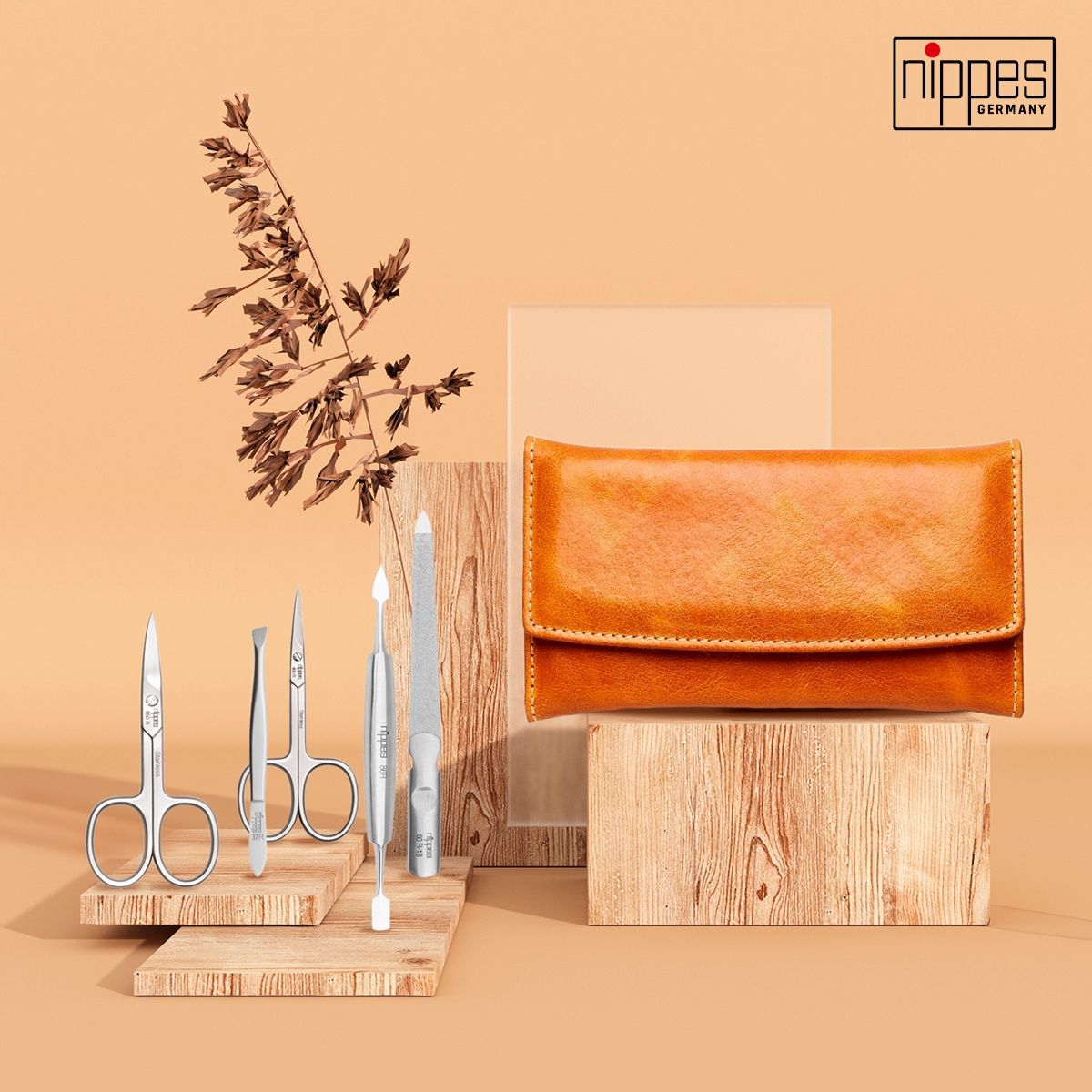 Maniküre-Etui COGNAC mit Maniküre-Instrumenten. Cognacfarbenes Lederetui. Logo 'nippes GERMANY'. Auf Holzblöcken arrangiert.