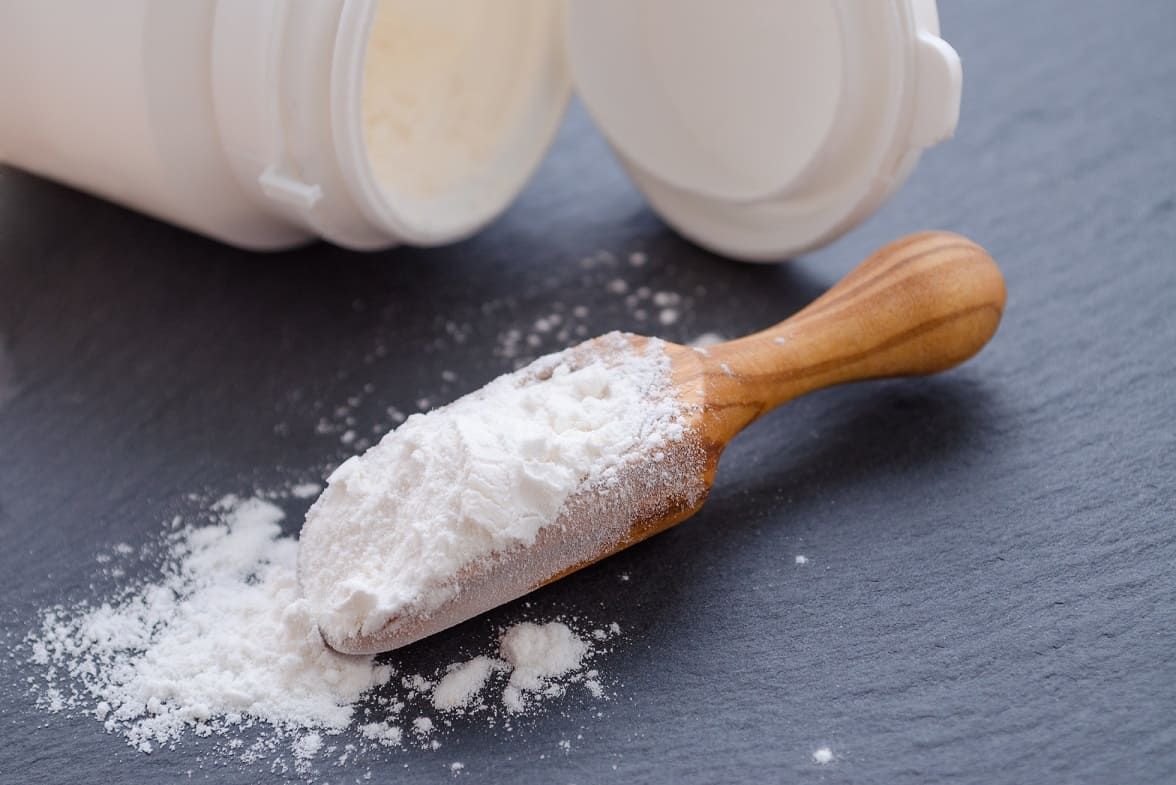 Weißes Pulver auf einem Holzlöffel. Daneben eine geöffnete weiße Dose. Auf dunklem Untergrund.