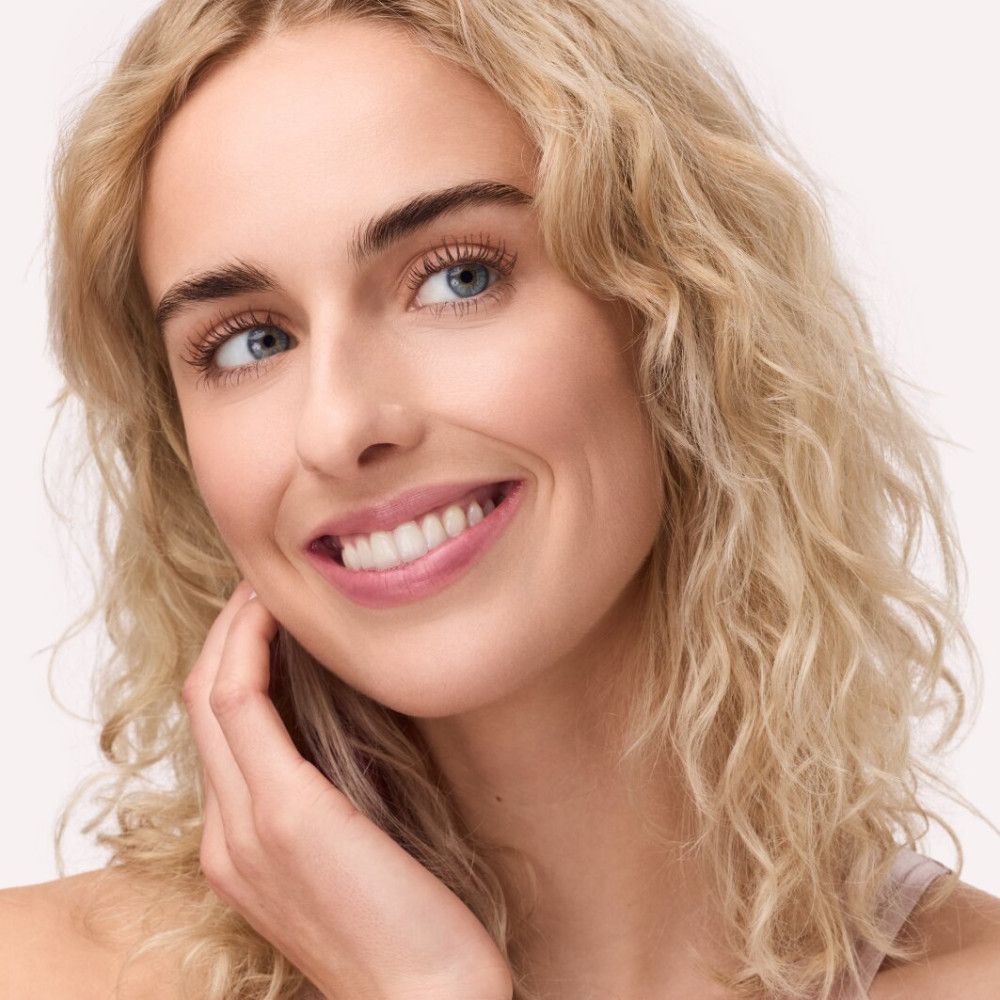 Porträt einer Frau mit blonden Locken und blauen Augen. Sie lächelt und berührt ihre Wange mit der Hand.