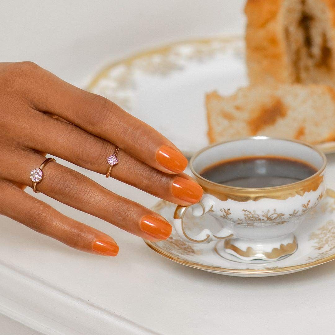 Hand mit lackierten, orangefarbenen Nägeln. Ringe. Kaffeetasse. Hintergrund: Gebäck.