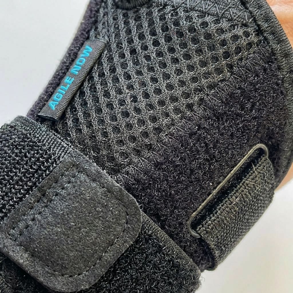 Nahaufnahme einer schwarzen Daumenbandage. Mesh-Gewebe, Klettverschlüsse und das Logo AGILE NOW sind sichtbar.