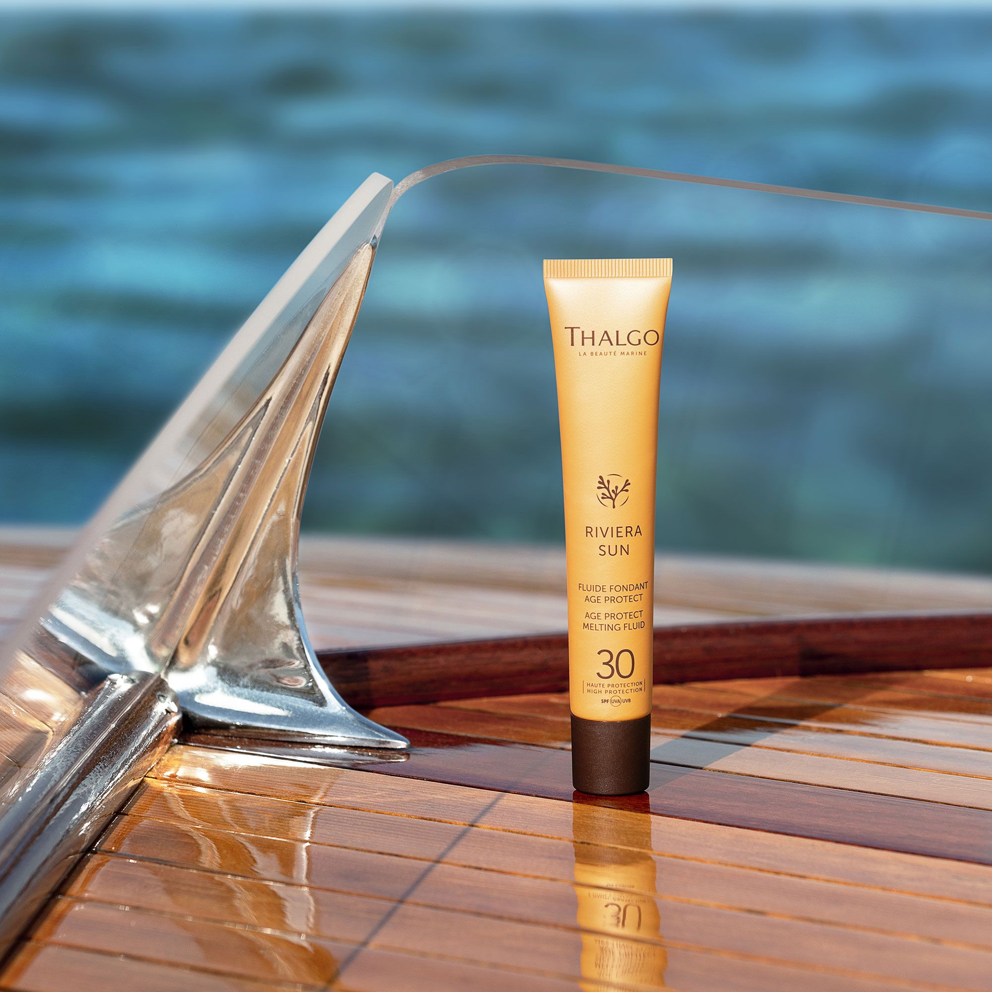 Gelbe Tube THALGO Riviera Sun Sonnenschutz-Fluid LSF 30 auf Holzoberfläche. Im Hintergrund Wasser und Bootsdetails.