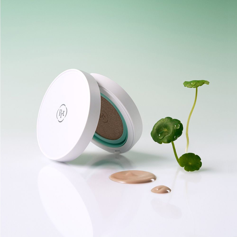 Geöffnete PURITO Centella BB Cushion-Verpackung mit Produkt und Tropfen daneben. Pflanze im Hintergrund.