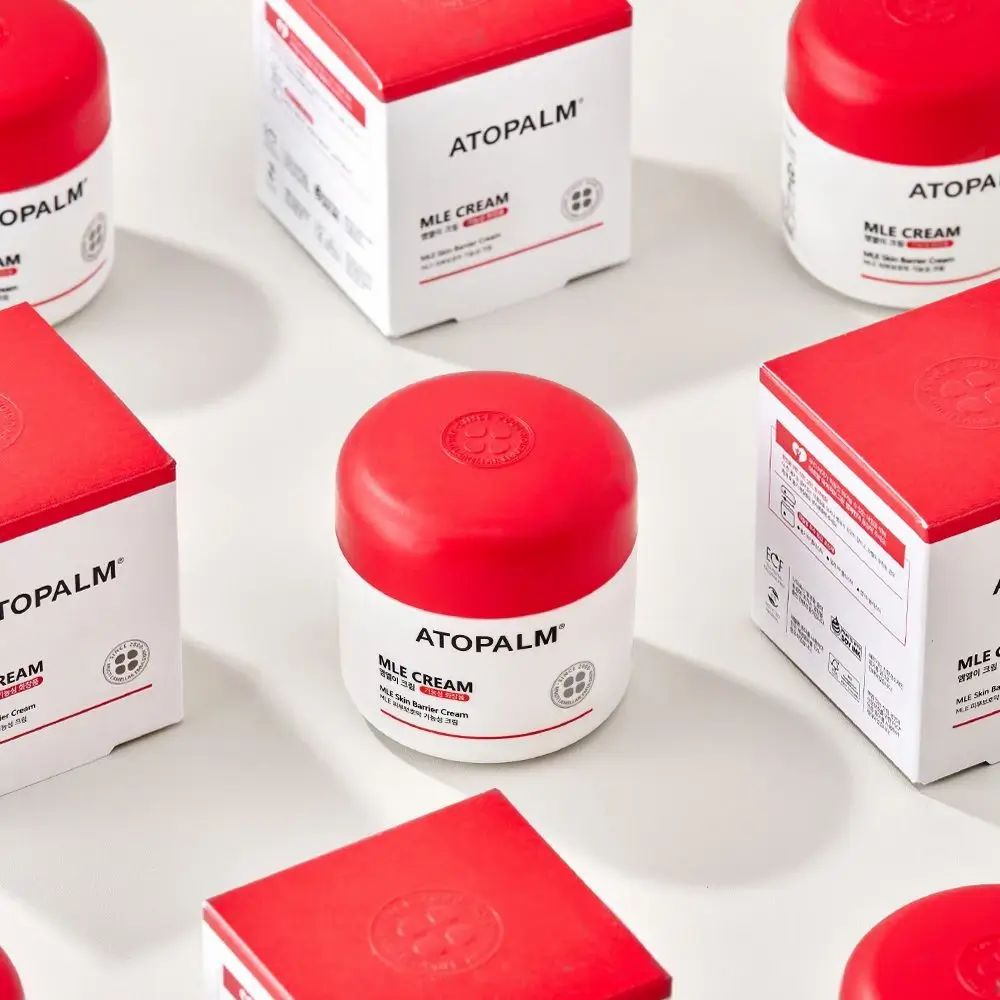 Mehrere quadratische Schachteln und ein Tiegel. Rote Deckel, weiße Seiten. Aufschrift: ATOPALM, MLE CREAM.