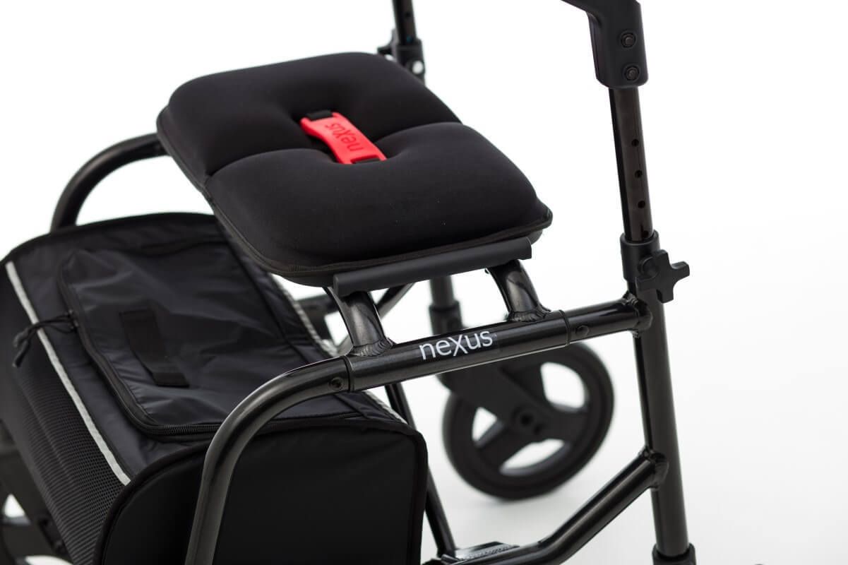 Nahaufnahme des Rollator-Sitzes mit Nexus-Logo. Schwarzer Sitz mit rotem Gurt. Einkaufstasche darunter.