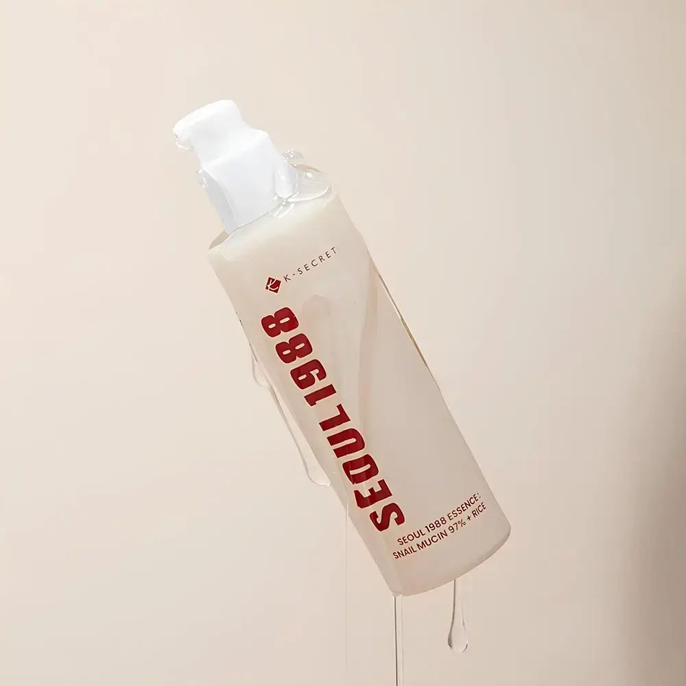 Beige Flasche mit weißem Pumpverschluss. Roter Schriftzug: SEOUL 1988. Flüssigkeit fließt. Text: K-SECRET, SEOUL 1988 ESSENCE, SNAIL MUCIN 97% + RICE.