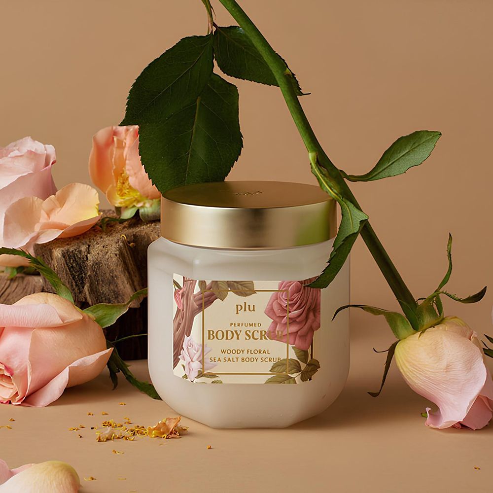 Glasbehälter mit goldfarbenem Deckel, umgeben von Rosen. Etikett: PLU Perfumed Body Scrub, Woody Floral Sea Salt Body Scrub.
