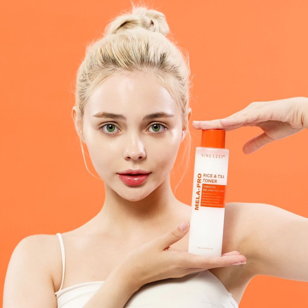 Eine Frau hält eine Flasche NINE LESS Mela-Pro Rice & TXA Toner. Orangefarbener Hintergrund. Die Frau blickt in die Kamera.