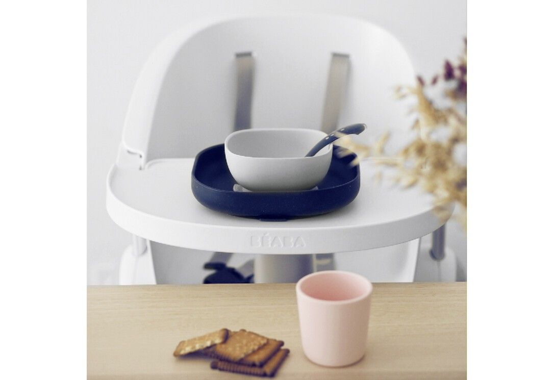 Vierteiliges BEABA-Geschirr-Set. Teller, Schüssel, Becher und Löffel. Auf einem Hochstuhl platziert. Rosa Becher, Kekse.