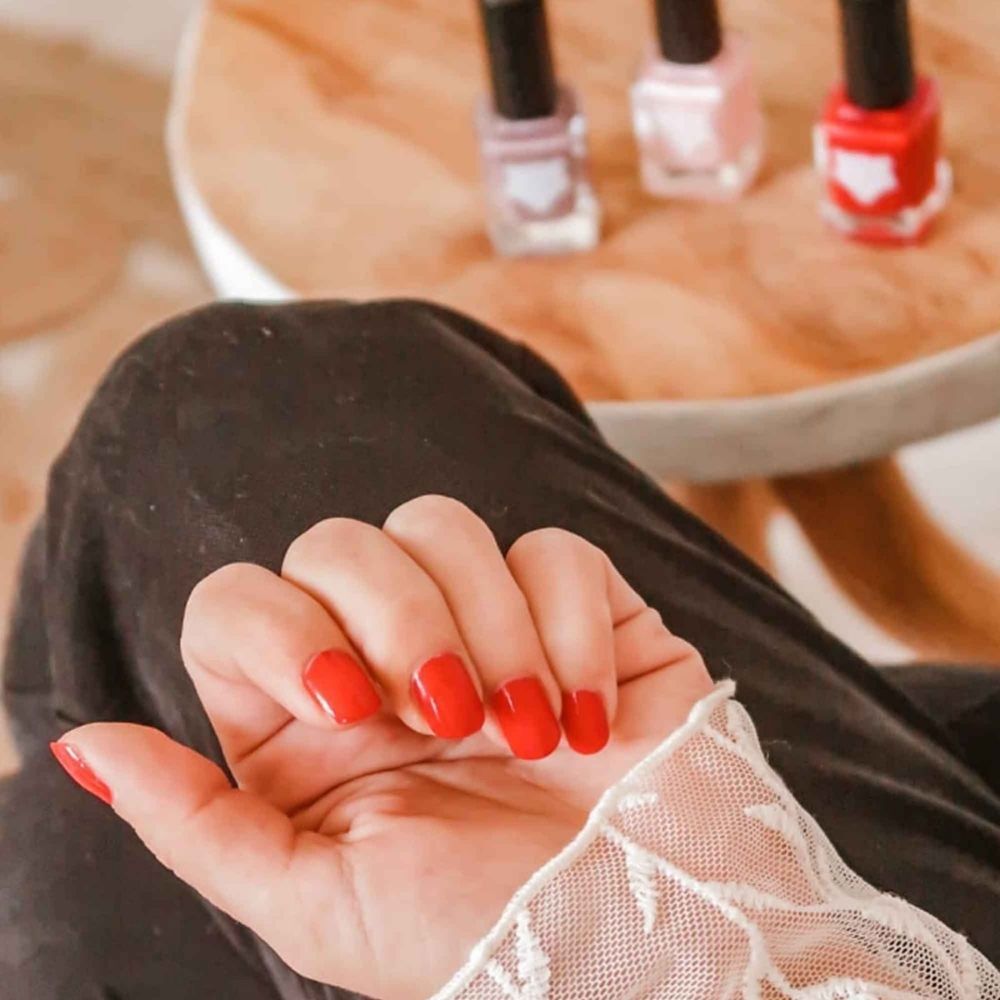 Rote Nägel einer Hand. Im Hintergrund stehen drei Nagellackflaschen auf einem kleinen Tisch.
