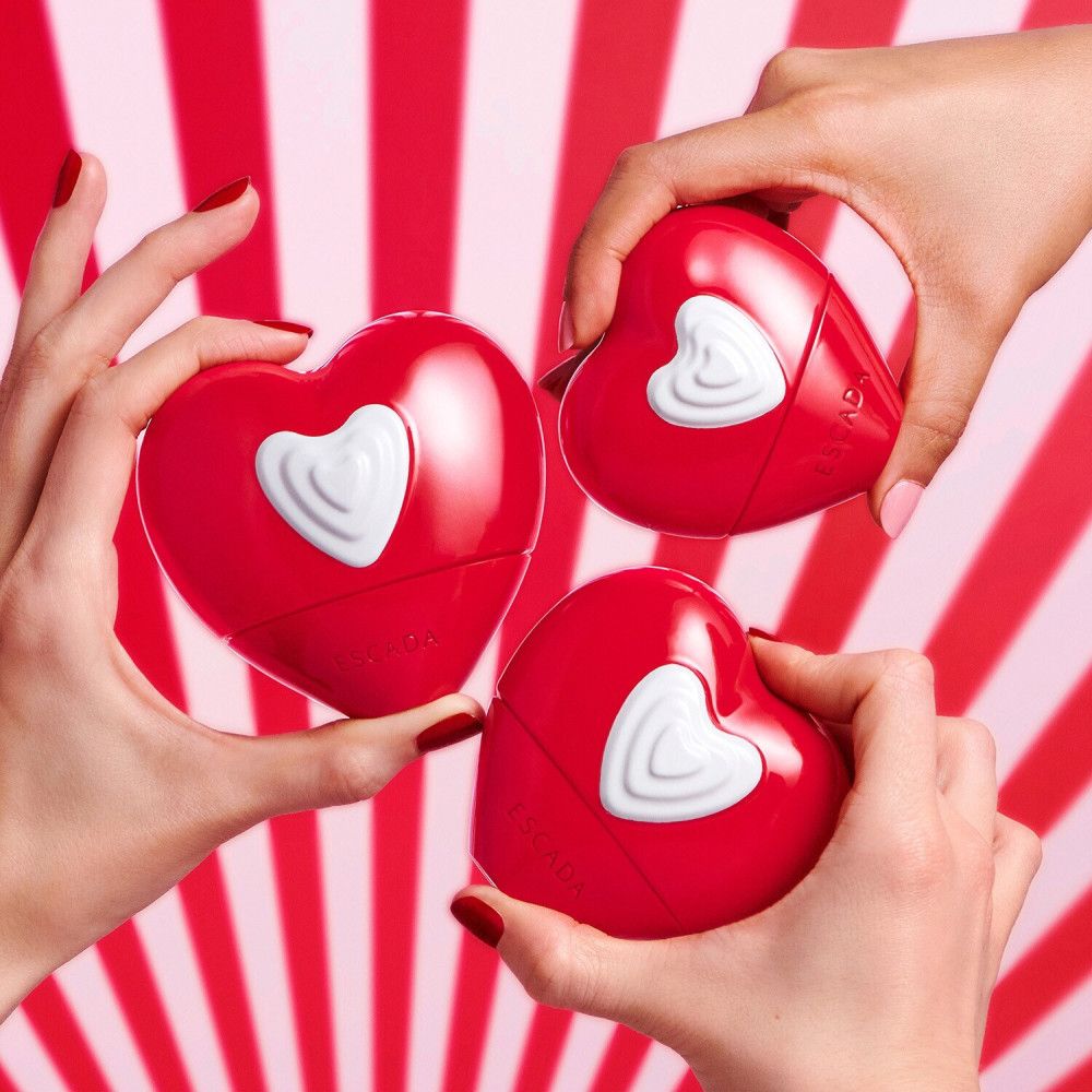 Drei rote Herzflakons mit weißem Herz, gehalten von Händen. Hintergrund mit roten und weißen Streifen.