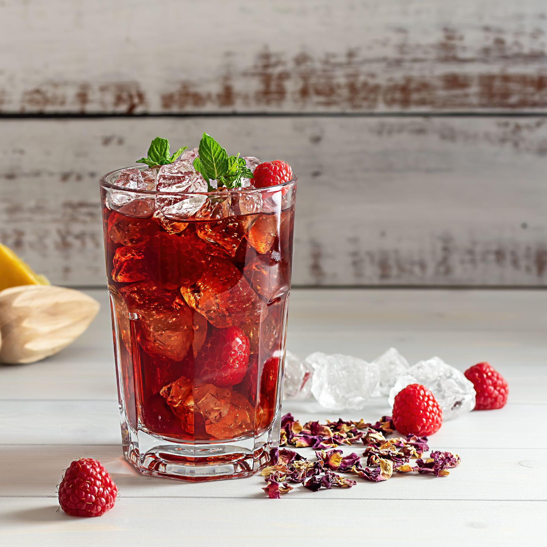 Glas mit rotem Getränk, Eiswürfeln, Himbeeren und Minze. Auf weißem Tisch. Streuung von Blütenblättern und Eiswürfeln.
